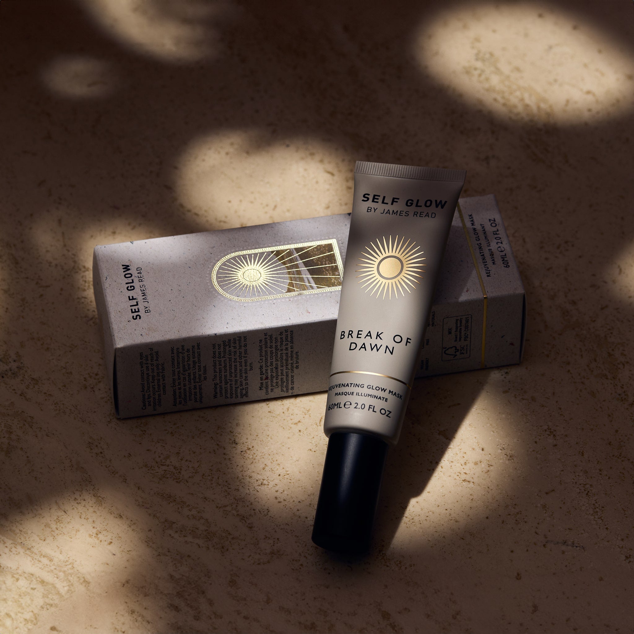 Self Glow by James Read Break of Dawn Rejuvenating Glow Mask bottle and box in a lifestyle image with dappled light, showcasing the product's radiance-boosting properties.