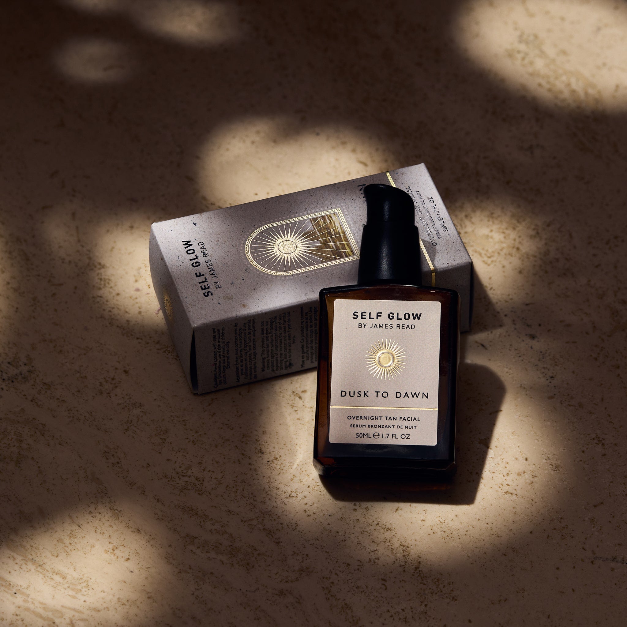 Self Glow by James Read Dusk to Dawn Overnight Tan Facial recyclable glass bottle leaning against a recycled cardboard box with vegetable-based inks and eco-friendly packaging, showcased under dappled light.