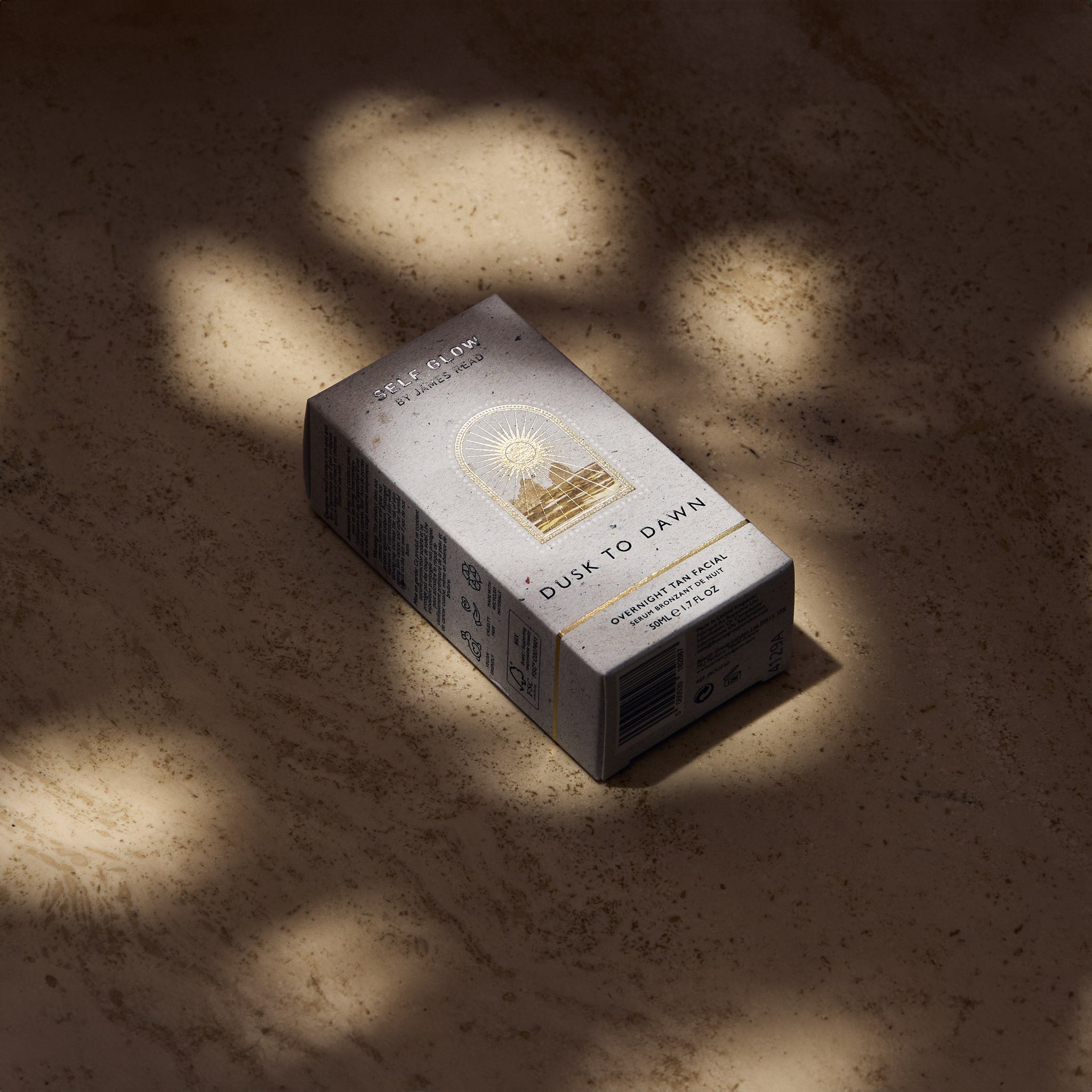 The recycled cardboard packaging box of Self Glow by James Read Dusk to Dawn Overnight Tan Facial, featuring vegetable-based inks and eco-friendly materials, displayed under dappled light.