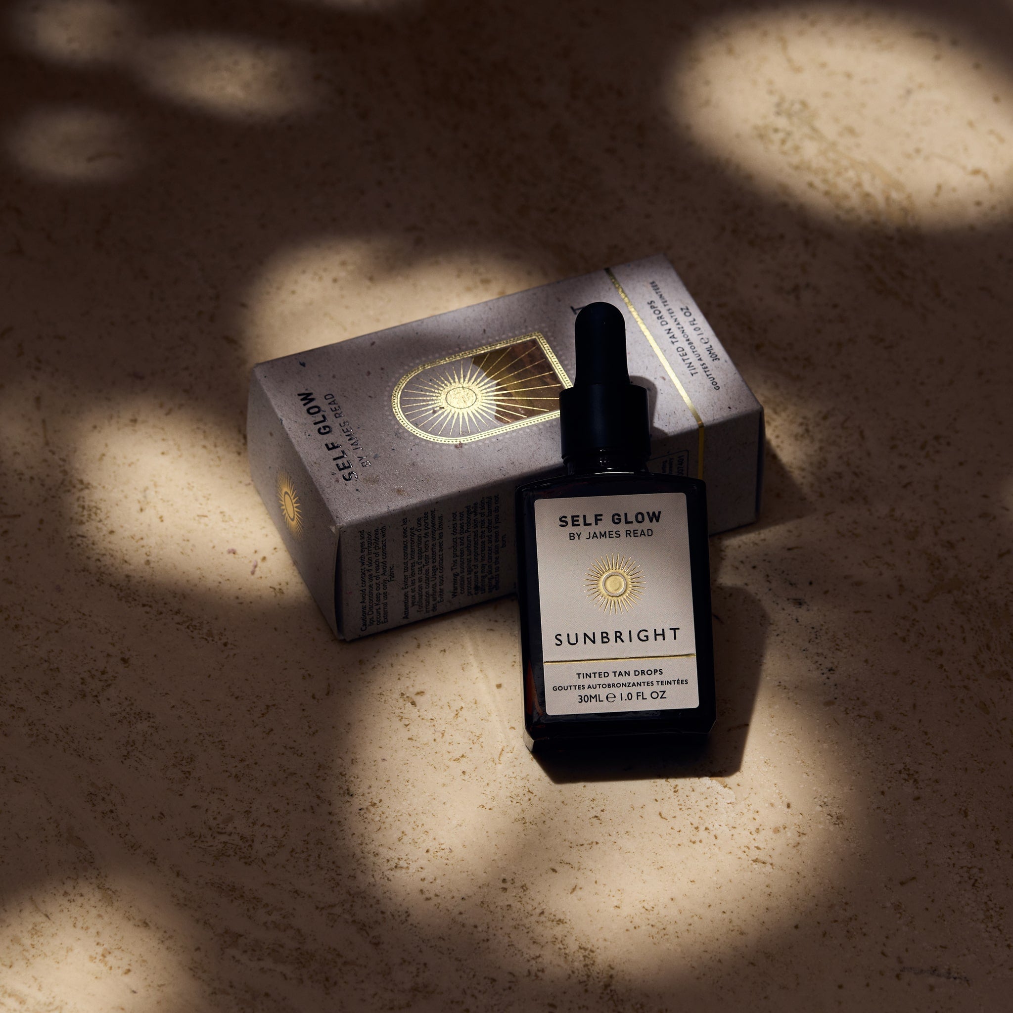 Self Glow by James Read Sunbright Tinted Tan Drops recyclable glass bottle leaning against a recycled cardboard box with vegetable-based inks and eco-friendly packaging, showcased under dappled light.