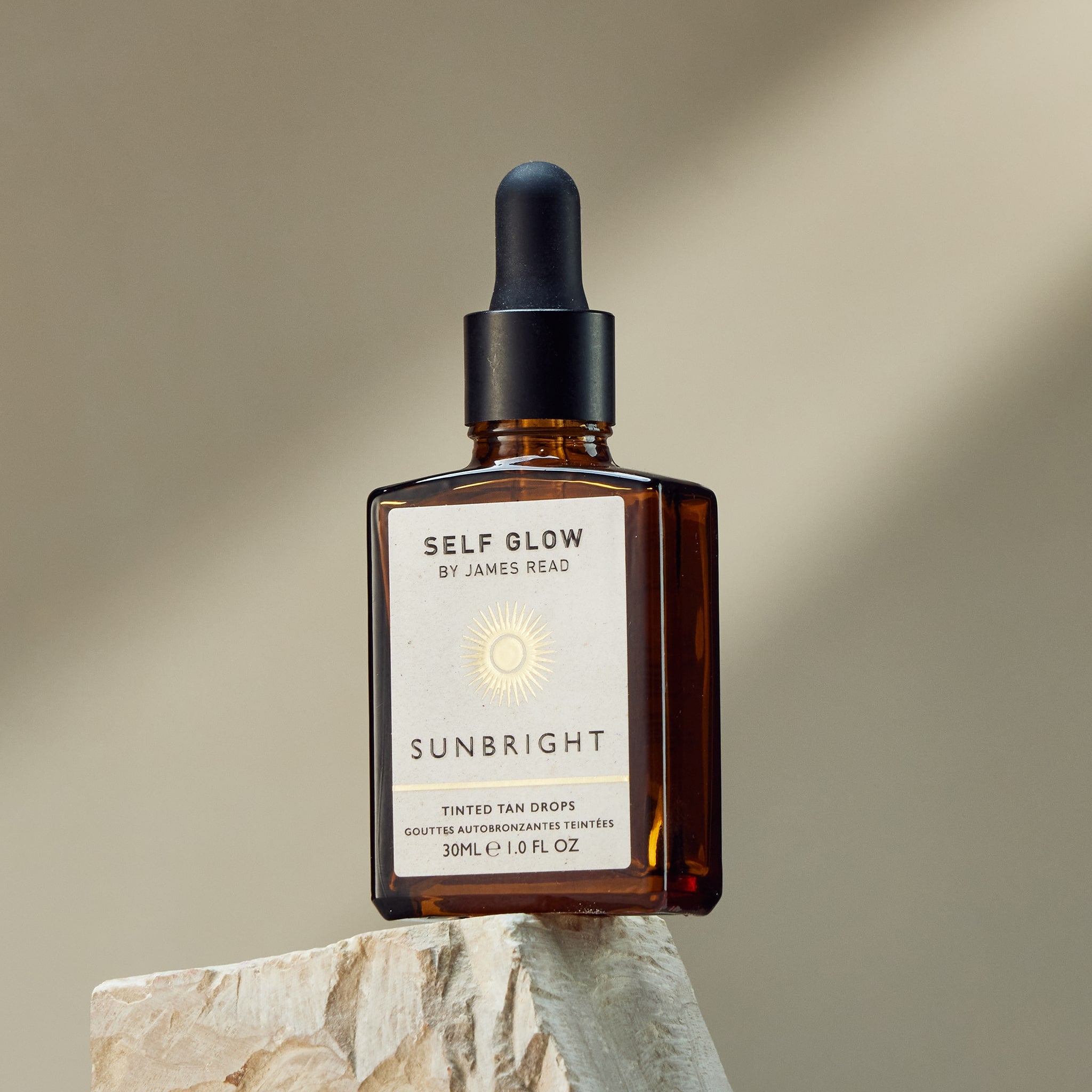 Self Glow by James Read Sunbright Tinted Tan Drops bottle in a lifestyle shot on a rock with the sun shining down, creating a shadow and emphasizing the product's ability to deliver a customizable, sun-kissed glow.