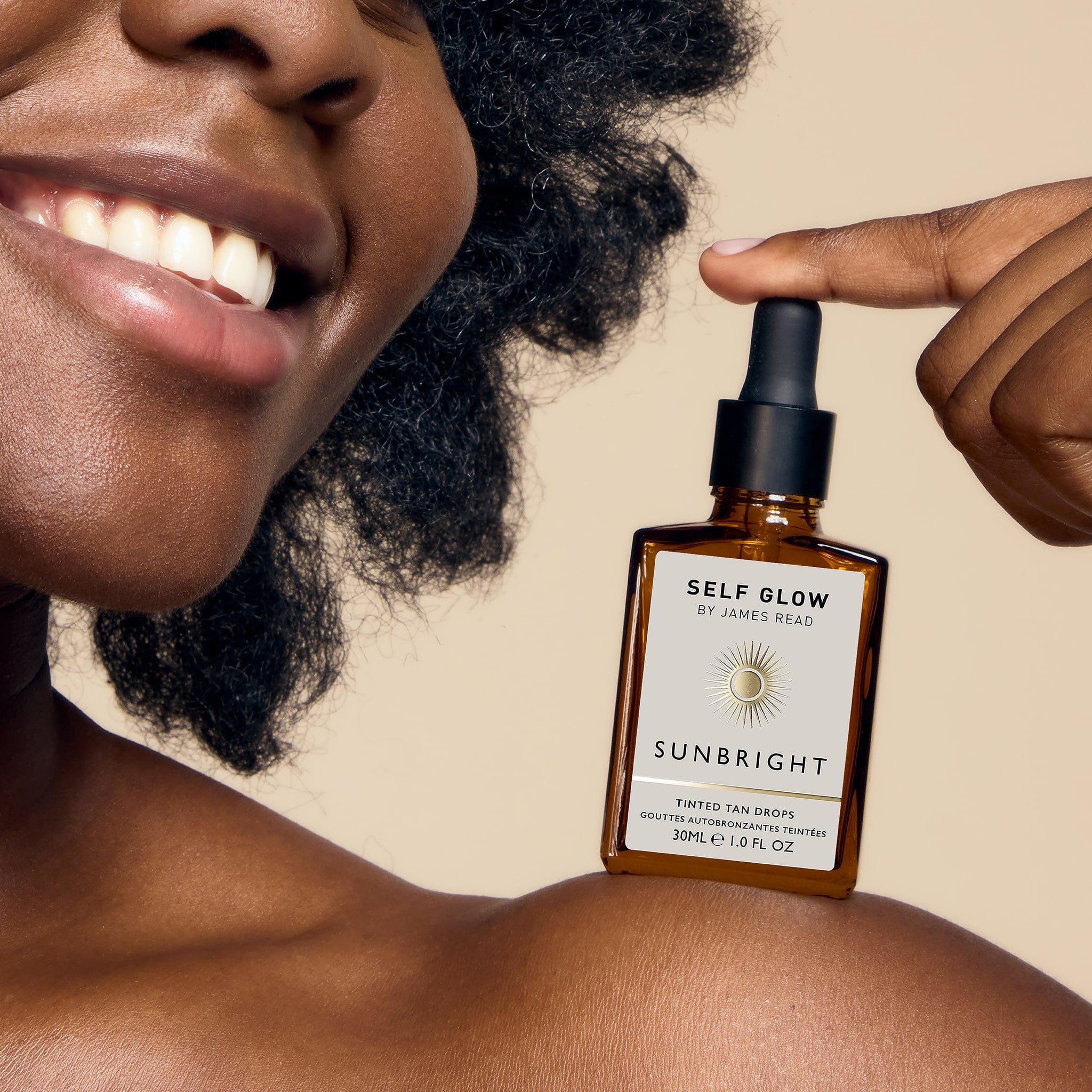 A black woman smiling and looking radiant with a glowing complexion, showcasing Self Glow by James Read Sunbright Tinted Tan Drops on her shoulder, highlighting the product's ability to seamlessly blend with moisturizer, serum, or makeup for instant results.