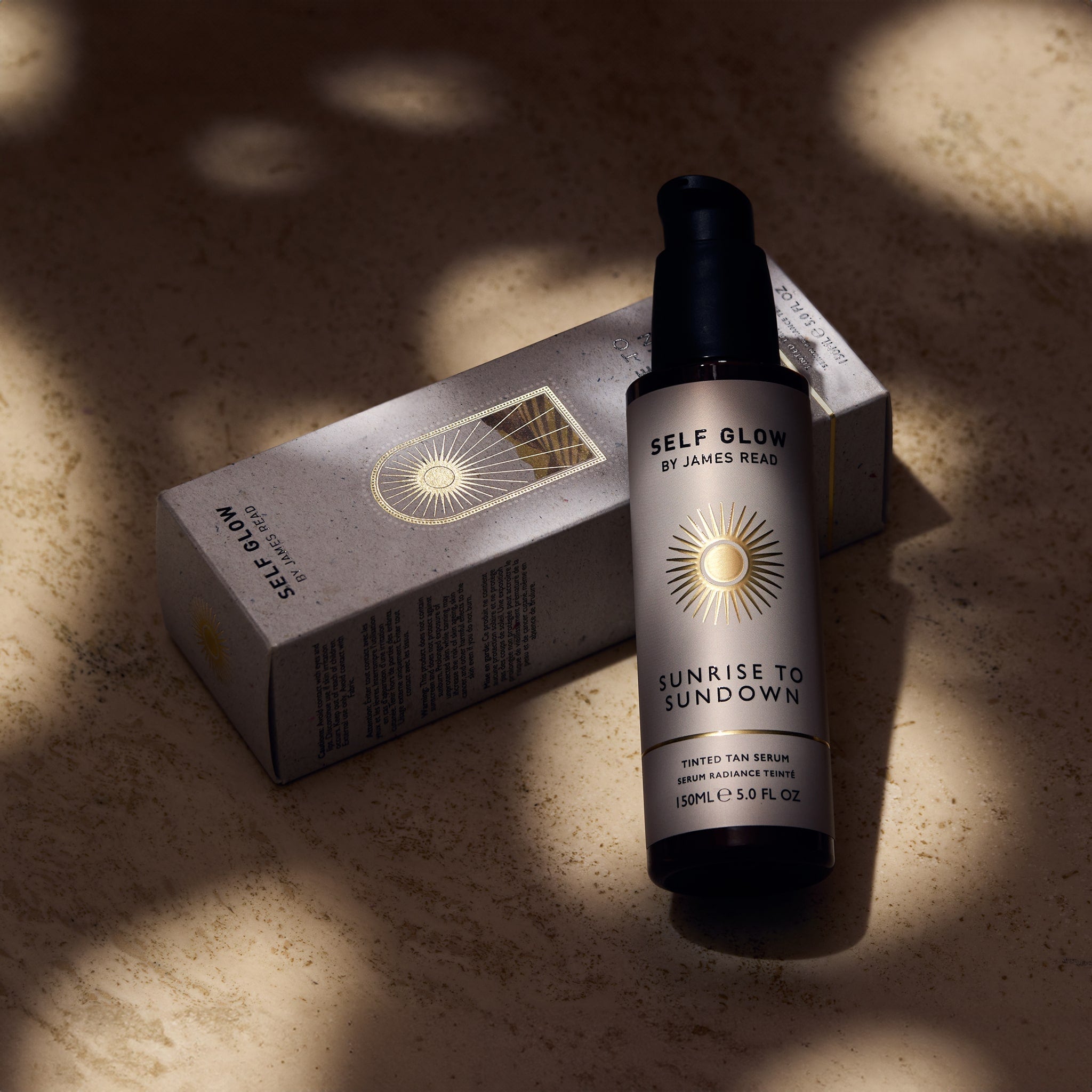 Self Glow by James Read Sunrise to Sundown Tinted Tan Serum recyclable glass bottle leaning against a recycled cardboard box with vegetable-based inks and eco-friendly packaging, showcased under dappled light.